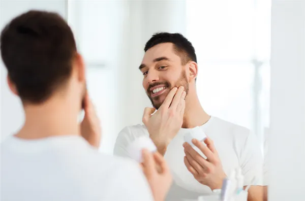 мужчина с короткой бородкой наносит перед зеркалом крем на лицо